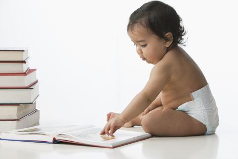 A baby sitting near a stack of books plays with the pages of an open book.