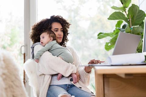 Una joven madre sostiene a su bebé sobre su hombro y busca información de salud en su computadora portátil.